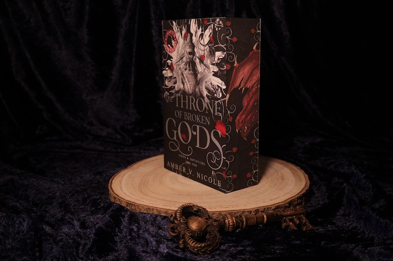 Peut inclure: Un livre intitul&eacute; "Throne of Broken Gods" d'Amber V. Nicole, avec une couverture noire orn&eacute;e de d&eacute;tails rouges et blancs. Le livre est pr&eacute;sent&eacute; sur une tranche de bois, avec une cl&eacute; d&eacute;corative devant. Roman fantastique.