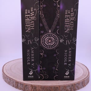 Peut inclure: Trois livres &agrave; couverture rigide avec des couvertures sombres, avec le titre "The Wrath of the Fallen" et le nom de l'auteur, Amber V. Nicole. Les dos sont noirs avec des accents violets et des motifs d&eacute;coratifs. Les livres sont pr&eacute;sent&eacute;s sur une tranche de bois.