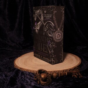 Peut inclure: Un livre &agrave; couverture rigide noir, intitul&eacute; "Wrath & Ruin: Gods & Monsters Book Four" par Amber V. Nicole, avec des d&eacute;tails argent&eacute;s et violets. Le livre est pr&eacute;sent&eacute; sur une tranche de bois, avec une cl&eacute; d&eacute;corative.