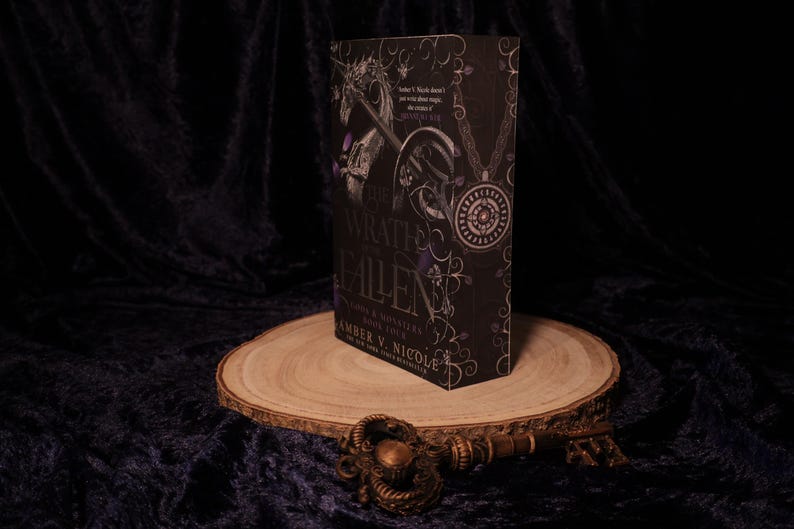 Peut inclure: Un livre &agrave; couverture rigide noir, intitul&eacute; "Wrath & Ruin: Gods & Monsters Book Four" par Amber V. Nicole, avec des d&eacute;tails argent&eacute;s et violets. Le livre est pr&eacute;sent&eacute; sur une tranche de bois, avec une cl&eacute; d&eacute;corative.