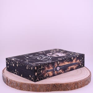 Peut inclure: Une bo&icirc;te noire avec des motifs floraux dor&eacute;s et le titre "The Book of Azrael" est pr&eacute;sent&eacute;e sur une tranche de bois. La bo&icirc;te pr&eacute;sente une &eacute;p&eacute;e et un &eacute;clair. Le nom de l'auteur, Amber V. Nicole, est &eacute;galement visible.