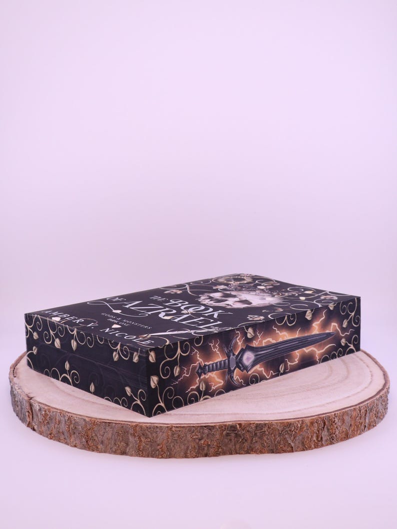 Peut inclure: Une bo&icirc;te noire avec des motifs floraux dor&eacute;s et le titre "The Book of Azrael" est pr&eacute;sent&eacute;e sur une tranche de bois. La bo&icirc;te pr&eacute;sente une &eacute;p&eacute;e et un &eacute;clair. Le nom de l'auteur, Amber V. Nicole, est &eacute;galement visible.