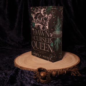Peut inclure: Un livre intitul&eacute; "Dawn of the Cursed Queen" est pos&eacute; sur une tranche de bois. Le livre a une couverture noire avec des accents argent&eacute;s et verts, et le titre est bien en &eacute;vidence. Une cl&eacute; d&eacute;corative repose devant le livre.