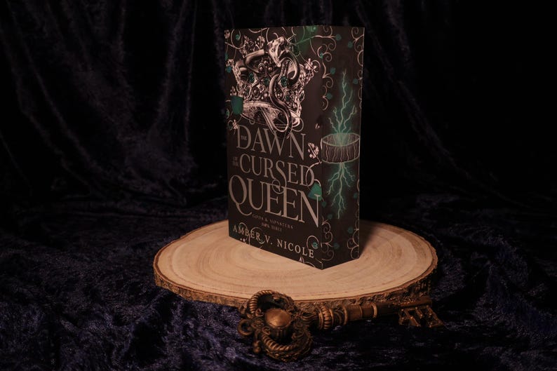 Peut inclure: Un livre intitul&eacute; "Dawn of the Cursed Queen" est pos&eacute; sur une tranche de bois. Le livre a une couverture noire avec des accents argent&eacute;s et verts, et le titre est bien en &eacute;vidence. Une cl&eacute; d&eacute;corative repose devant le livre.