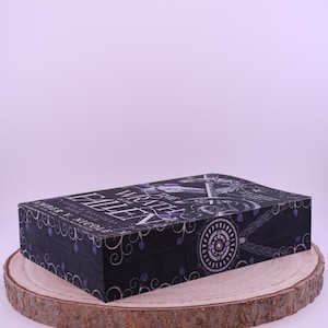Peut inclure: Un coffret de livre noir avec le titre "Wrath of the Fallen" en lettres blanches et violettes. La bo&icirc;te pr&eacute;sente un motif d&eacute;coratif avec des motifs floraux et circulaires. La bo&icirc;te repose sur une tranche de bois ronde.