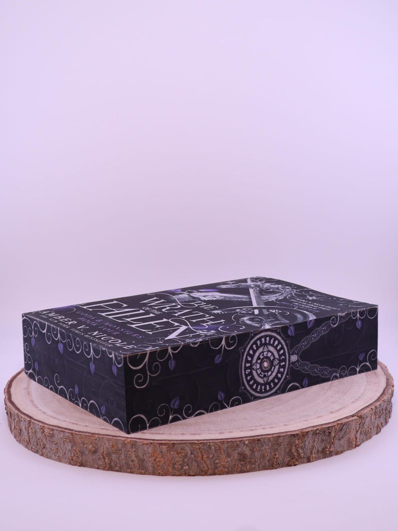 Peut inclure: Un coffret de livre noir avec le titre "Wrath of the Fallen" en lettres blanches et violettes. La bo&icirc;te pr&eacute;sente un motif d&eacute;coratif avec des motifs floraux et circulaires. La bo&icirc;te repose sur une tranche de bois ronde.