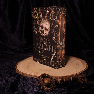 Peut inclure: Un livre &agrave; couverture rigide intitul&eacute; "The Book of Azrael" avec un cr&acirc;ne et une &eacute;p&eacute;e sur la couverture. Le livre est pr&eacute;sent&eacute; sur une tranche de bois avec une cl&eacute; d&eacute;corative au premier plan. La couverture est noire avec des accents dor&eacute;s et orange.