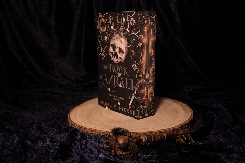 Peut inclure: Un livre &agrave; couverture rigide intitul&eacute; "The Book of Azrael" avec un cr&acirc;ne et une &eacute;p&eacute;e sur la couverture. Le livre est pr&eacute;sent&eacute; sur une tranche de bois avec une cl&eacute; d&eacute;corative au premier plan. La couverture est noire avec des accents dor&eacute;s et orange.