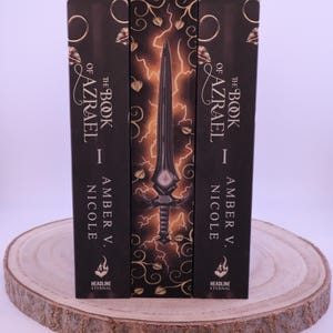 Peut inclure: Trois livres &agrave; couverture rigide intitul&eacute;s "The Book of Azrael I" par Amber V. Nicole. Les dos sont noirs avec des lettres dor&eacute;es et des &eacute;l&eacute;ments d&eacute;coratifs. Le livre central pr&eacute;sente un dessin d'&eacute;p&eacute;e avec des &eacute;clairs.