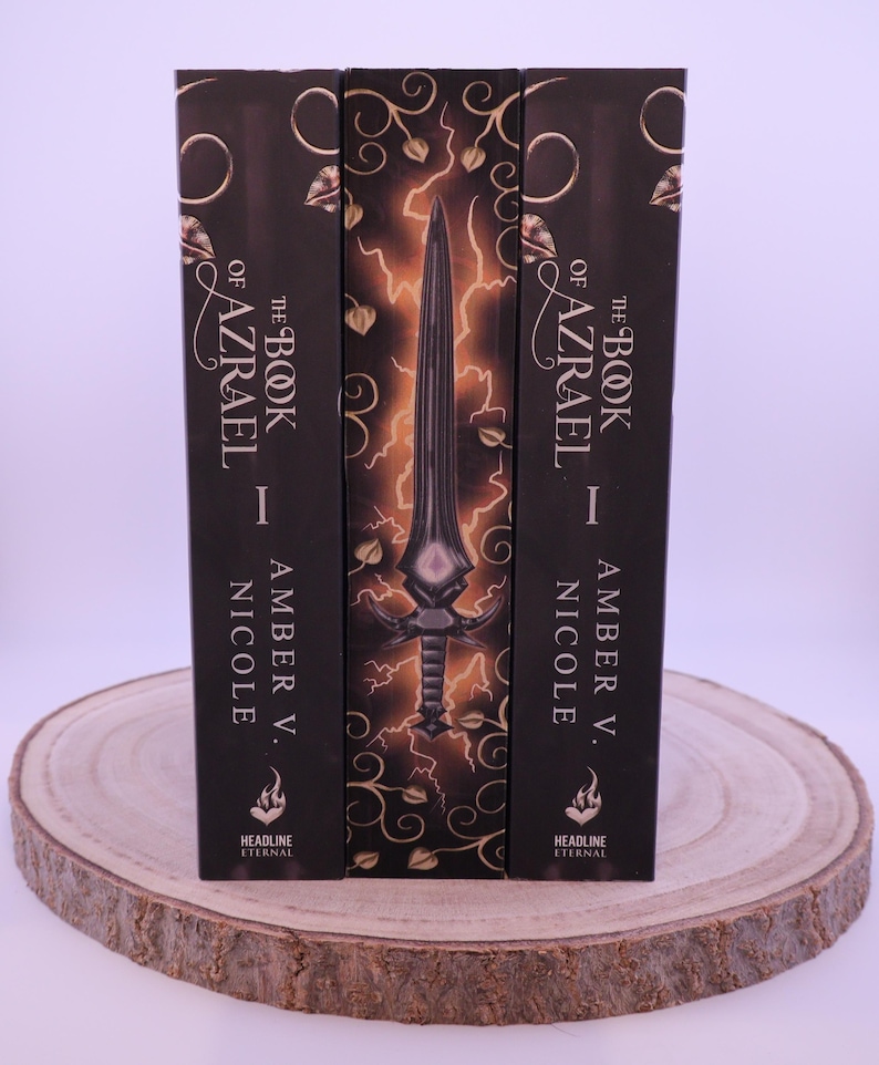 Peut inclure: Trois livres &agrave; couverture rigide intitul&eacute;s "The Book of Azrael I" par Amber V. Nicole. Les dos sont noirs avec des lettres dor&eacute;es et des &eacute;l&eacute;ments d&eacute;coratifs. Le livre central pr&eacute;sente un dessin d'&eacute;p&eacute;e avec des &eacute;clairs.