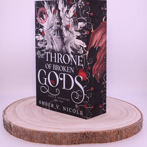 Peut inclure: Un livre &agrave; couverture rigide intitul&eacute; "The Throne of Broken Gods" par Amber V. Nicole. Le livre a une couverture noire avec du texte blanc et une illustration d&eacute;taill&eacute;e d'un dragon et d'autres &eacute;l&eacute;ments fantastiques. Le livre est pr&eacute;sent&eacute; sur une tranche de bois.