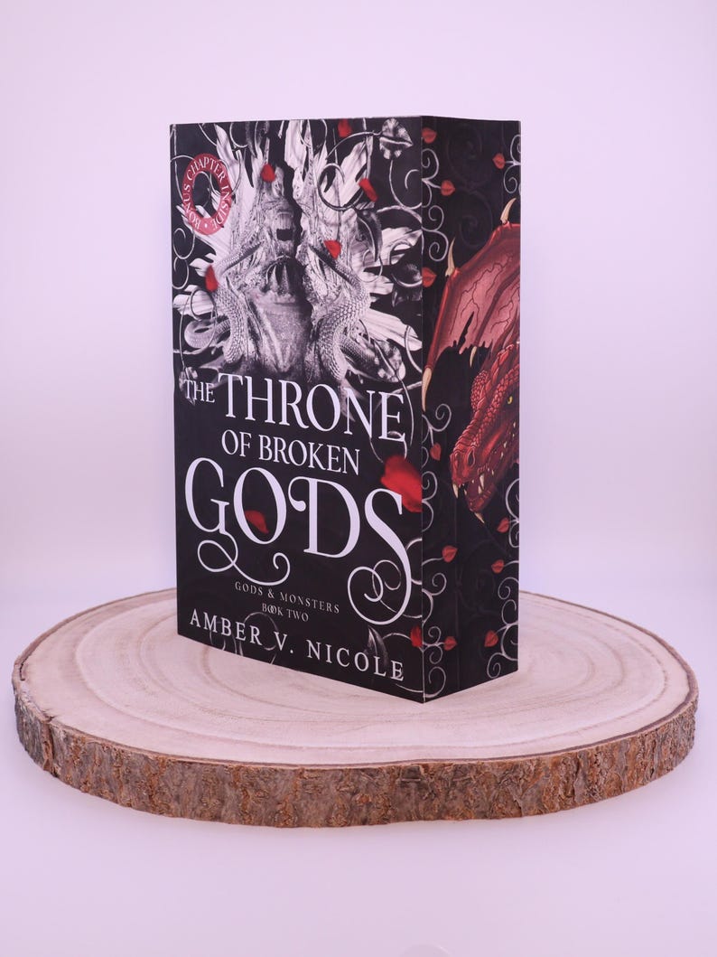 Peut inclure: Un livre &agrave; couverture rigide intitul&eacute; "The Throne of Broken Gods" par Amber V. Nicole. Le livre a une couverture noire avec du texte blanc et une illustration d&eacute;taill&eacute;e d'un dragon et d'autres &eacute;l&eacute;ments fantastiques. Le livre est pr&eacute;sent&eacute; sur une tranche de bois.