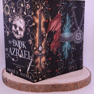 Peut inclure: Une pile de livres de fantasy, "The Book of Azrael" d'Amber V. Nicole. Les couvertures pr&eacute;sentent des illustrations d'un cr&acirc;ne, d'une &eacute;p&eacute;e, d'un dragon et d'un pendentif sur fond noir avec des motifs floraux. Les livres sont pr&eacute;sent&eacute;s sur une tranche de bois.