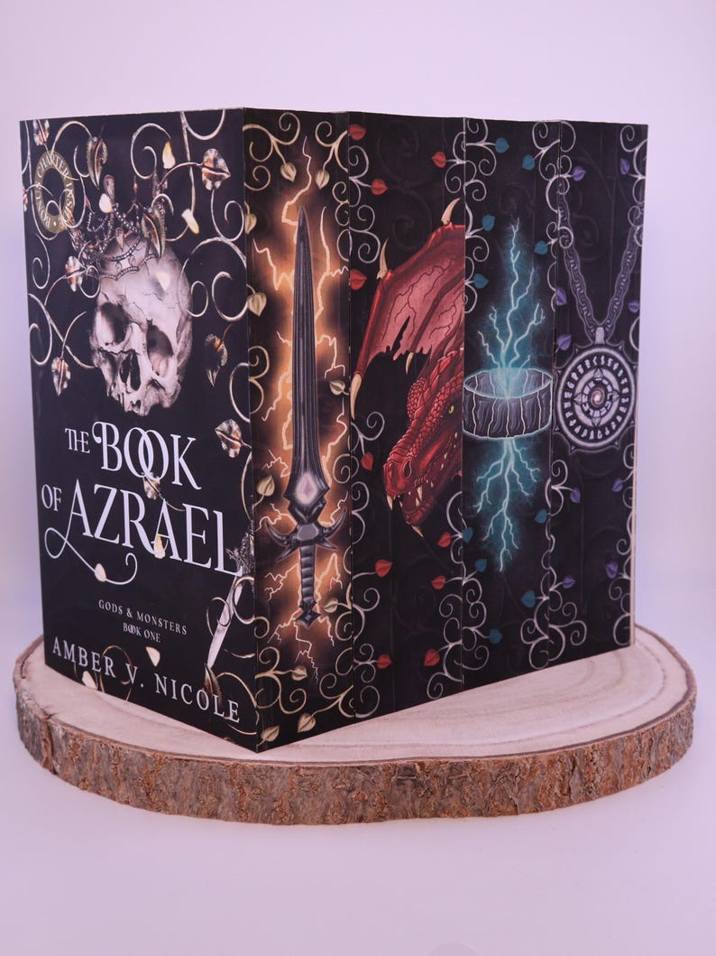 Peut inclure: Une pile de livres de fantasy, "The Book of Azrael" d'Amber V. Nicole. Les couvertures pr&eacute;sentent des illustrations d'un cr&acirc;ne, d'une &eacute;p&eacute;e, d'un dragon et d'un pendentif sur fond noir avec des motifs floraux. Les livres sont pr&eacute;sent&eacute;s sur une tranche de bois.