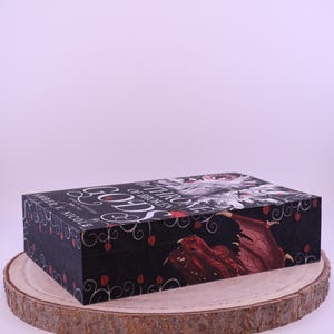 Peut inclure: Bo&icirc;te rectangulaire noire avec un th&egrave;me fantastique, avec une illustration de dragon rouge et le texte "Gods of Broken Throne". La bo&icirc;te est d&eacute;cor&eacute;e d'accents floraux rouges et de texte blanc. Repose sur une tranche de bois.