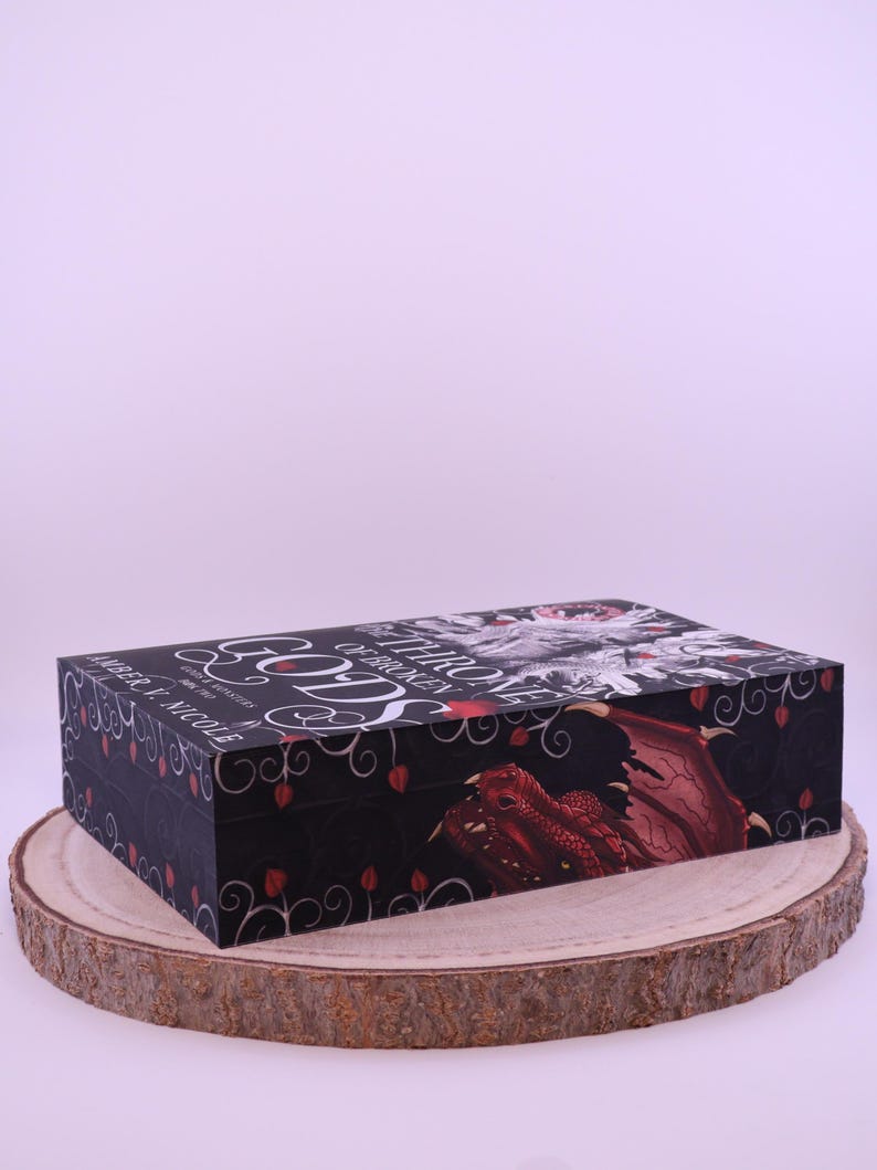 Peut inclure: Bo&icirc;te rectangulaire noire avec un th&egrave;me fantastique, avec une illustration de dragon rouge et le texte "Gods of Broken Throne". La bo&icirc;te est d&eacute;cor&eacute;e d'accents floraux rouges et de texte blanc. Repose sur une tranche de bois.