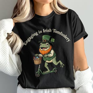 以下が含まれることがあります： 「Engaging in Irish Tomfoolery」の文字が入った黒いTシャツ。イラストは、レプラコーンの帽子とベストを着てスクーターに乗り、卵のバスケットを持っているカエルです。カエルは赤いひげを生やしています。