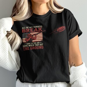May include: Black t-shirt with a graphic featuring a shooter aiming a rifle, an American flag, and a flying clay target. Text reads "ALL OF MY TARGETS BREAK...SOME IN THE AIR SOME WHEN THEY HIT THE GROUND."