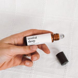 Könnte beinhalten: Eine kleine, braune ätherische Öl-Rollerflasche mit einem weißen Etikett mit der Aufschrift "restful sleep". Die Flasche wird in einer Hand gehalten, mit abgenommener schwarzer Kappe, die daneben platziert ist. Der Hintergrund ist eine weiße, strukturierte Oberfläche.