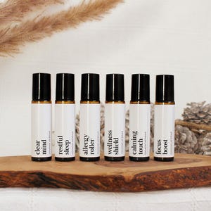 Könnte beinhalten: Sechs ätherische Öl-Rollerflaschen mit schwarzen Kappen und weißen Etiketten sind auf einem Holzbrett angeordnet. Die Etiketten lauten "clear mind", "restful sleep", "allergy roller", "wellness shield", "calming touch" und "focus boost". Dekorative getrocknete Pflanzen und Tannenzapfen sind im Hintergrund.