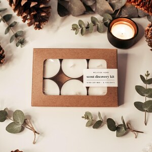 May include: A brown cardboard box with six white soy wax tealights. The box has a rectangular window revealing the candles. The label reads "MELVIDA HOME scent discovery kit". The image includes a lit candle in a black holder, pinecones, and eucalyptus.