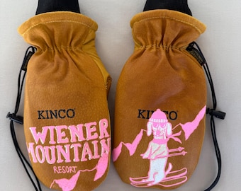 May include: A pair of brown leather mittens with black cuffs and drawstrings. The mittens feature pink graphics and text that read "KINCO WIENER MOUNTAIN RESORT" and a cartoon dog skiing. The mittens are designed for winter wear.
