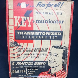 Könnte beinhalten: Vintage "KEY municator" Transistor-Telegrafie-Set-Box. Die Box ist rot, weiß und schwarz mit dem Text "Fun for all!" und "A Practical Hobby!". Enthält eine Illustration eines lächelnden Mannes mit Kopfhörern.