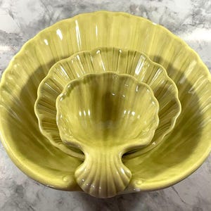 May include: A set of three yellow ceramic shell-shaped serving dishes. The largest dish is a shallow bowl, with two smaller dishes nested inside. The dishes have a ribbed texture and a glossy finish, perfect for serving appetizers or snacks.