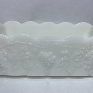 Pode incluir: Uma jardineira retangular de vidro leitoso branco com uma borda superior recortada e um padrão em relevo de cachos de uvas e folhas de videira que envolvem as laterais externas do recipiente de vidro opaco.