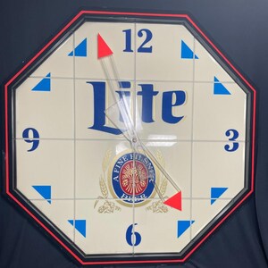 May include: An octagonal wall clock with a cream-colored face, featuring the word "Lite" in blue, and the numbers 12, 3, 6, and 9. The clock has a black and red border, with blue triangles and red-tipped hands.