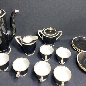 May include: A black and white porcelain tea set with gold trim. The set includes a teapot, sugar bowl, creamer, six cups, and saucers. The cups and saucers have a scalloped edge.