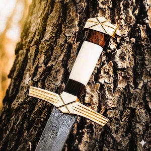 May include: A detailed image of a sword with a patterned blade and a handle featuring white, dark brown, and gold accents. The sword is leaning against a textured tree trunk, showcasing its craftsmanship and design.