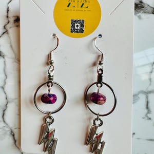 May include: A pair of silver earrings with a lightning bolt charm. Each earring features a purple and pink stone bead suspended within a silver hoop. The earrings hang from a silver hook. The earrings are displayed on a white card with a yellow logo.