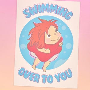K&ouml;nnte beinhalten: Ein Druck mit einer Zeichentrickfigur mit roten Haaren und einem roten Kleid, die in einem blauen Kreis mit Blasen schwebt. Der Text "SWIMMING OVER TO YOU" steht in Blau &uuml;ber und unter der Figur.
