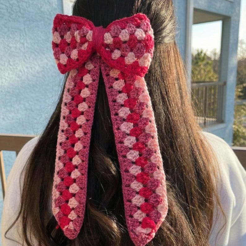 以下が含まれることがあります： ピンクと赤の色合いの、手作りのかぎ針編みヘアリボン。リボンは、長く垂れ下がったテールが付いた、大きく装飾的な形状をしています。かぎ針編みのパターンは、クラシックなグラニースクエアのデザインで、アクセサリーに質感と視覚的な面白さを加えています。