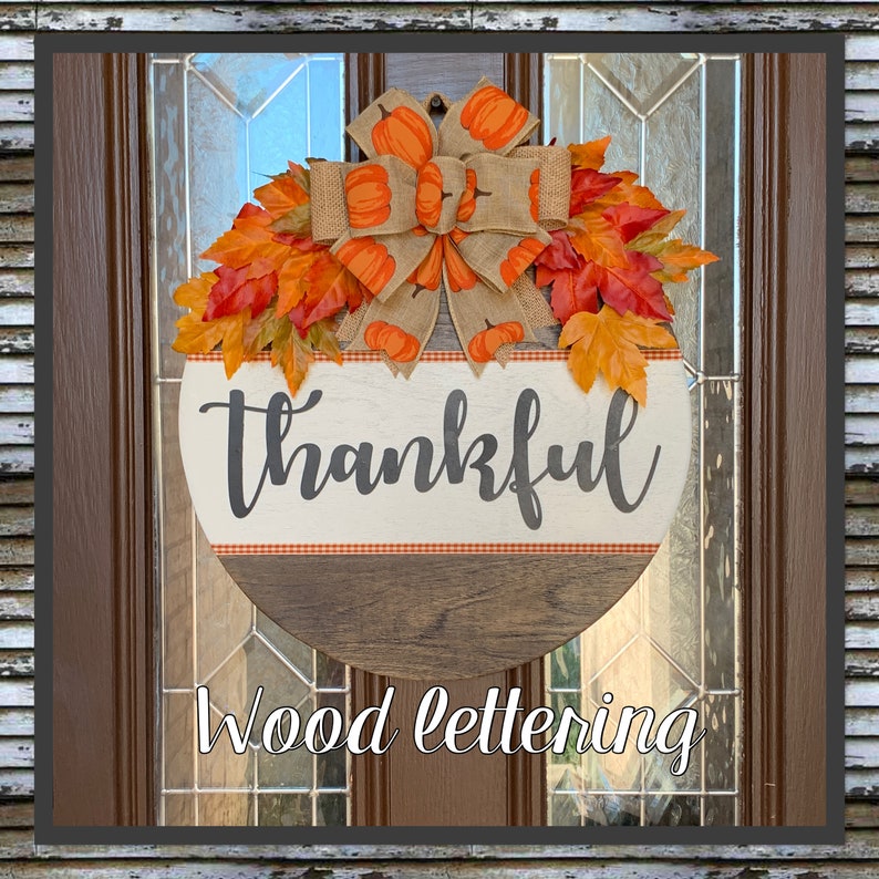 May include: A round wooden sign with the word 'thankful' written in gray cursive lettering on a white background with a red and white checkered border. The sign is decorated with a burlap bow and orange and brown fall leaves. The sign is hanging on a brown wooden door with a glass window. The text 'Wood lettering' is written below the sign.