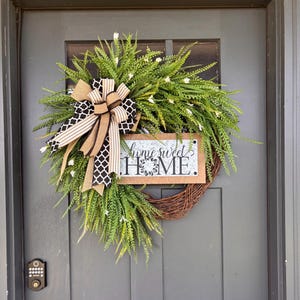 May include: A round grapevine wreath featuring lush green foliage with small white flowers. A decorative sign with a galvanized metal center and wood frame displays the text "home sweet HOME". A large bow made of burlap and black-and-white patterned ribbon.