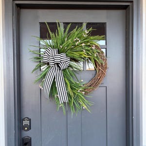 May include: A decorative wreath with a natural grapevine base, adorned with green foliage, small pink flowers, and a black and white striped bow. The wreath is hanging on a gray door.