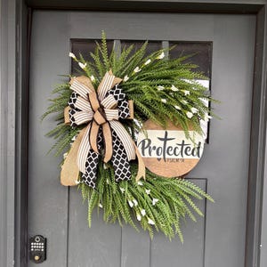 May include: A decorative wreath featuring lush green foliage with small white flowers, accented by a large bow made of tan burlap and black-and-white patterned ribbon. A round wooden sign is attached, displaying the text Protected and Psalm 91.