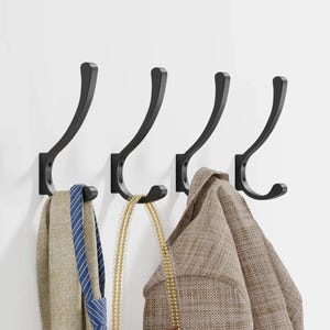 May include: Four black metal wall hooks with a curved design. A beige scarf with a blue striped tie, a gold beaded necklace, and a tan jacket hang from the hooks. The hooks are mounted on a white wall.
