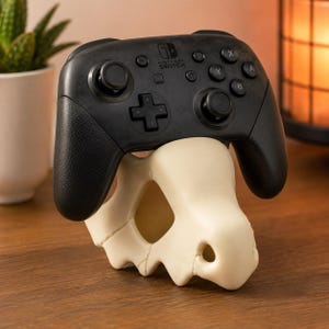 May include: A black Nintendo Switch Pro controller resting on a cream-colored, bone-shaped display stand. The controller features two analog sticks, a directional pad, and buttons labeled A, B, X, and Y. The stand has a hollow, organic structure.