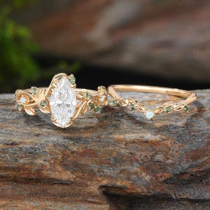 Pode incluir: Um conjunto de anéis de noivado dourados com um diamante central em corte marquise. O anel é adornado com detalhes de folhas e videiras, acentuados por pequenas pedras preciosas verdes e pedras opalescentes. O conjunto inclui uma aliança correspondente.