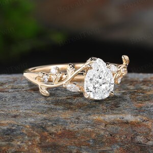 May include: A gold engagement ring with a pear-shaped diamond center stone. The ring features a delicate vine design with leaves and smaller accent diamonds. The ring is set on a natural stone surface.