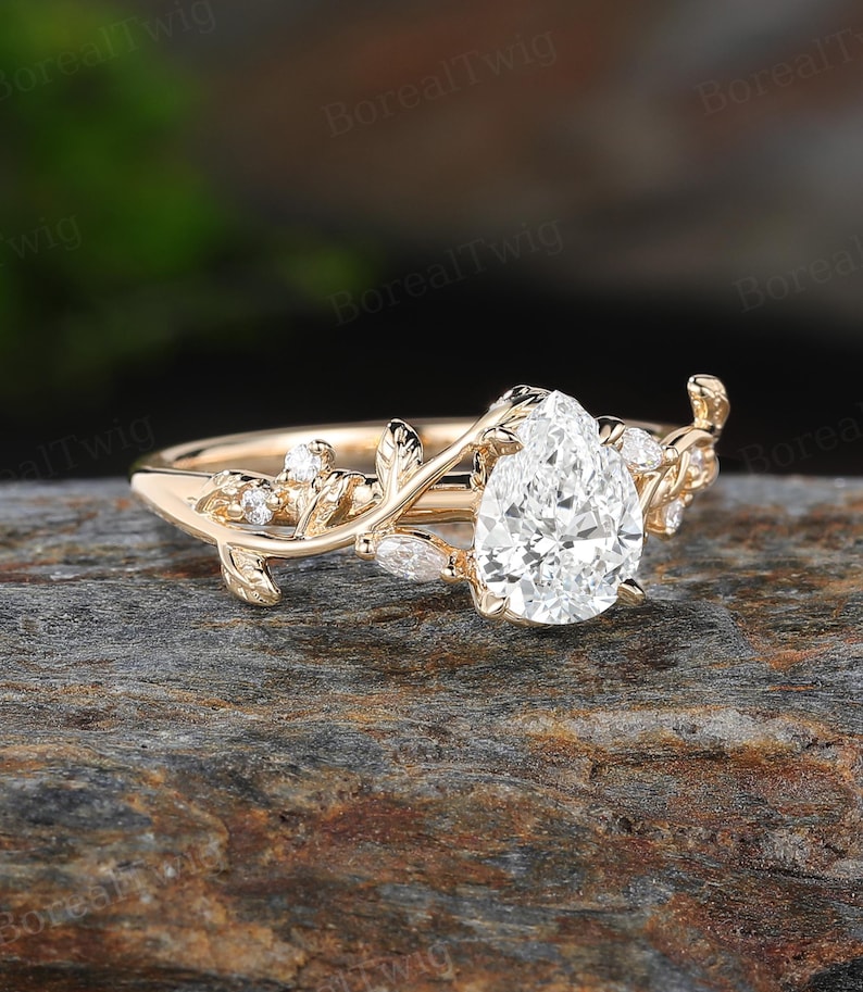 May include: A gold engagement ring with a pear-shaped diamond center stone. The ring features a delicate vine design with leaves and smaller accent diamonds. The ring is set on a natural stone surface.