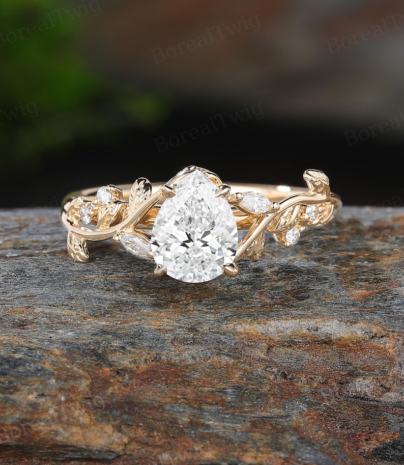 May include: A gold engagement ring with a pear-shaped diamond center stone. The ring features a delicate vine design with small accent diamonds and leaf details. The ring is set on a stone surface.