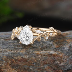 May include: A gold ring with a pear-shaped diamond center stone. The ring features a delicate leaf and vine design with small accent diamonds. The ring is set on a natural stone surface.