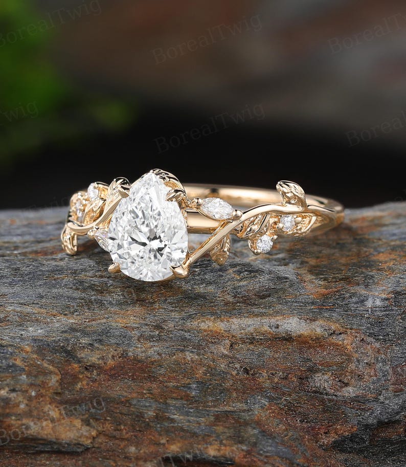 May include: A gold ring with a pear-shaped diamond center stone. The ring features a delicate leaf and vine design with small accent diamonds. The ring is set on a natural stone surface.