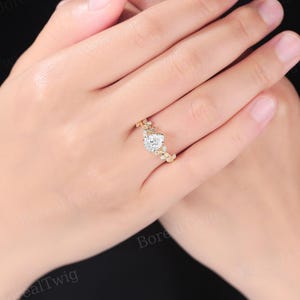 May include: A gold ring featuring a pear-shaped, clear gemstone as the centerpiece. The ring has a delicate, floral design with small, round gemstones accenting the band. The ring is set against a neutral background.