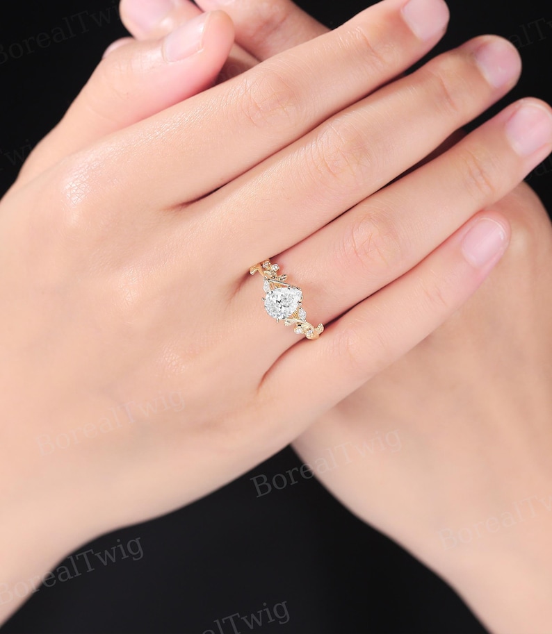 May include: A gold ring featuring a pear-shaped, clear gemstone as the centerpiece. The ring has a delicate, floral design with small, round gemstones accenting the band. The ring is set against a neutral background.