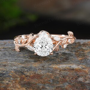 May include: Rose gold engagement ring featuring a pear-shaped diamond center stone. The band is adorned with delicate leaf and vine detailing, and small accent diamonds. The ring is set on a textured, dark gray surface.